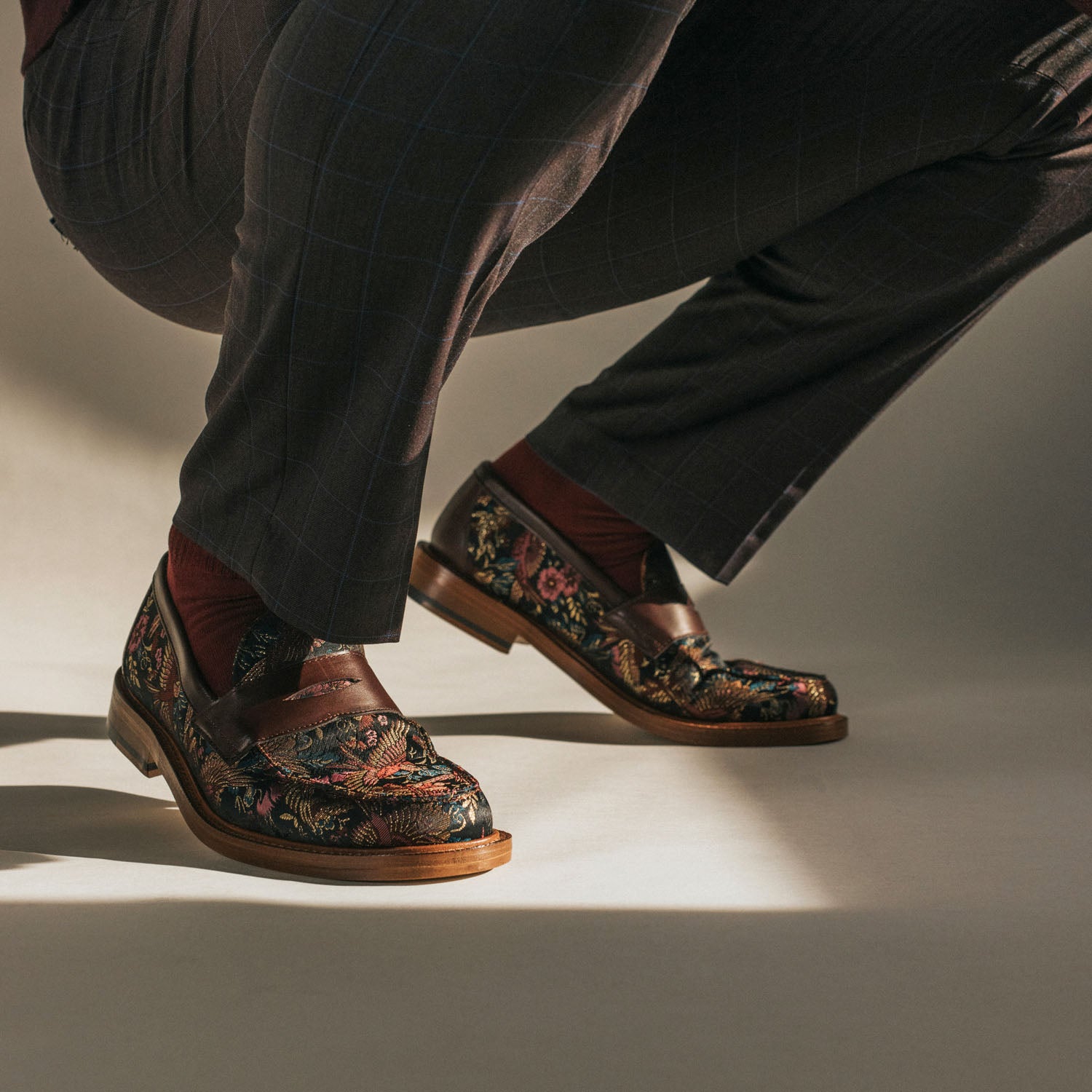 A person wears black pants and floral-patterned loafers with brown straps, squatting on the ground with one knee bent.