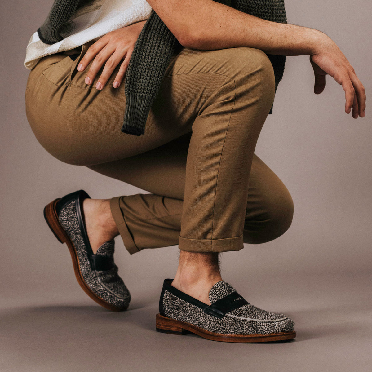 Person wearing tan pants, a white shirt, and a green cardigan squatting down, showcasing gray loafers with black details and wooden soles.