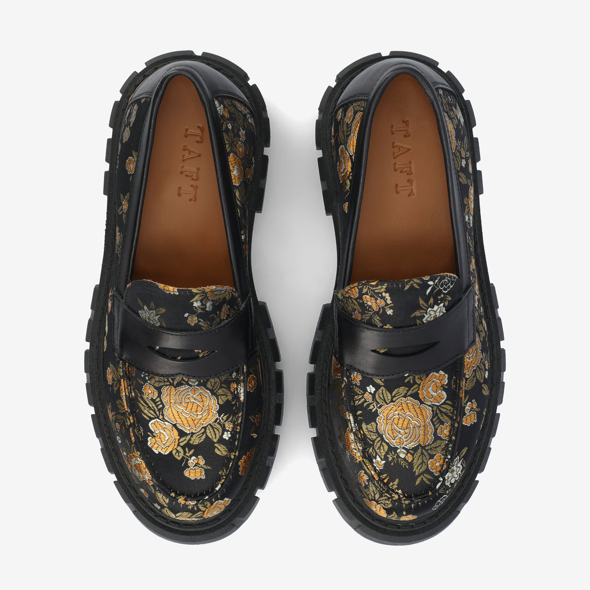 A pair of black floral loafers with rugged soles and brown leather insoles are displayed from a top-down perspective. They feature gold and white flower patterns.