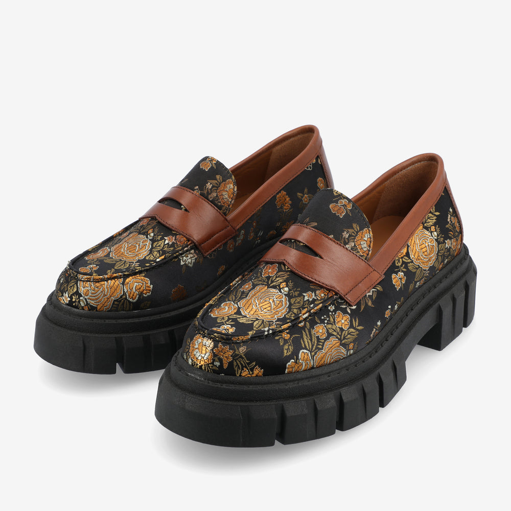 A pair of black loafers with chunky black soles and decorative yellow floral patterns, featuring brown leather straps across the top.