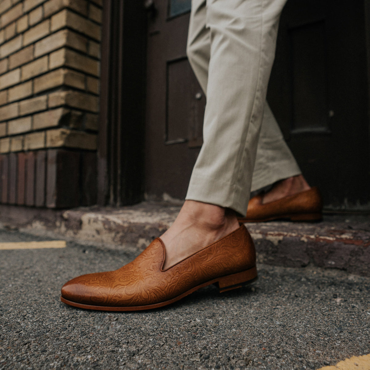 A person wearing light pants and patterned brown loafers stands on a concrete surface near a brick wall and a wooden door.