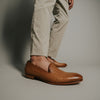 A person wearing beige pants and brown leather slip-on shoes with embossed patterns, standing against a neutral background.