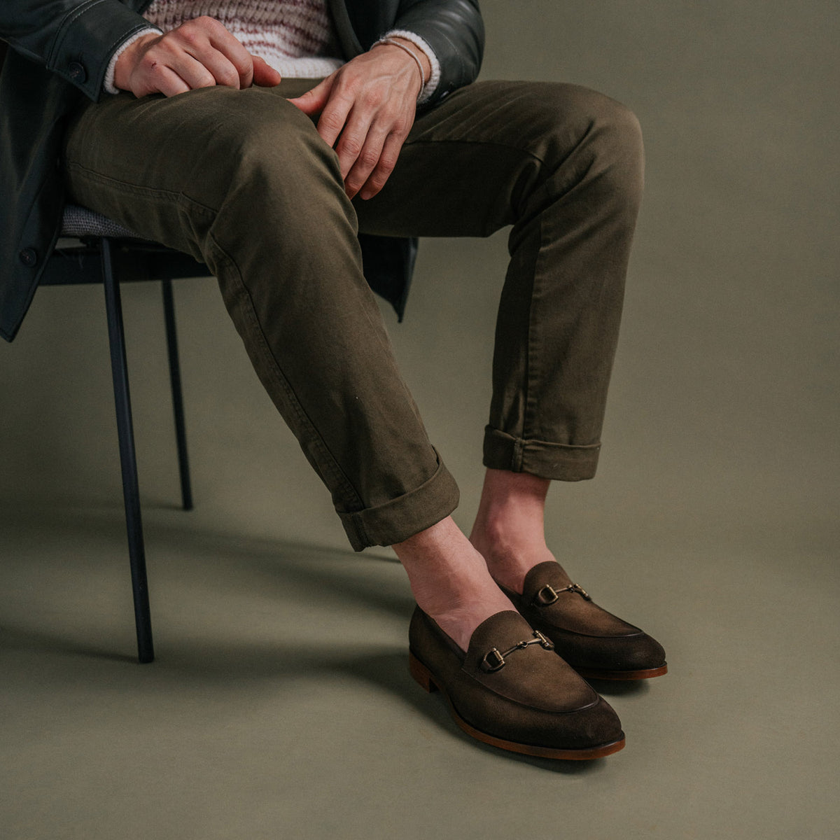 A person wearing olive green pants and brown loafers sits on a chair with their hands resting on their knees.