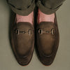 Close-up of a pair of feet wearing brown suede loafers with metal bit detailing, standing on a beige surface. The person is wearing dark pants.