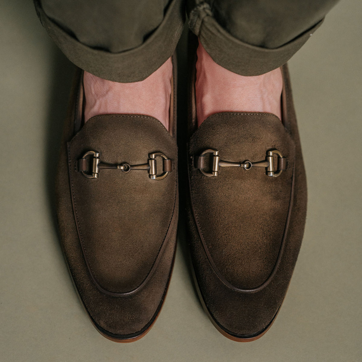 Close-up of a pair of feet wearing brown suede loafers with metal bit detailing, standing on a beige surface. The person is wearing dark pants.