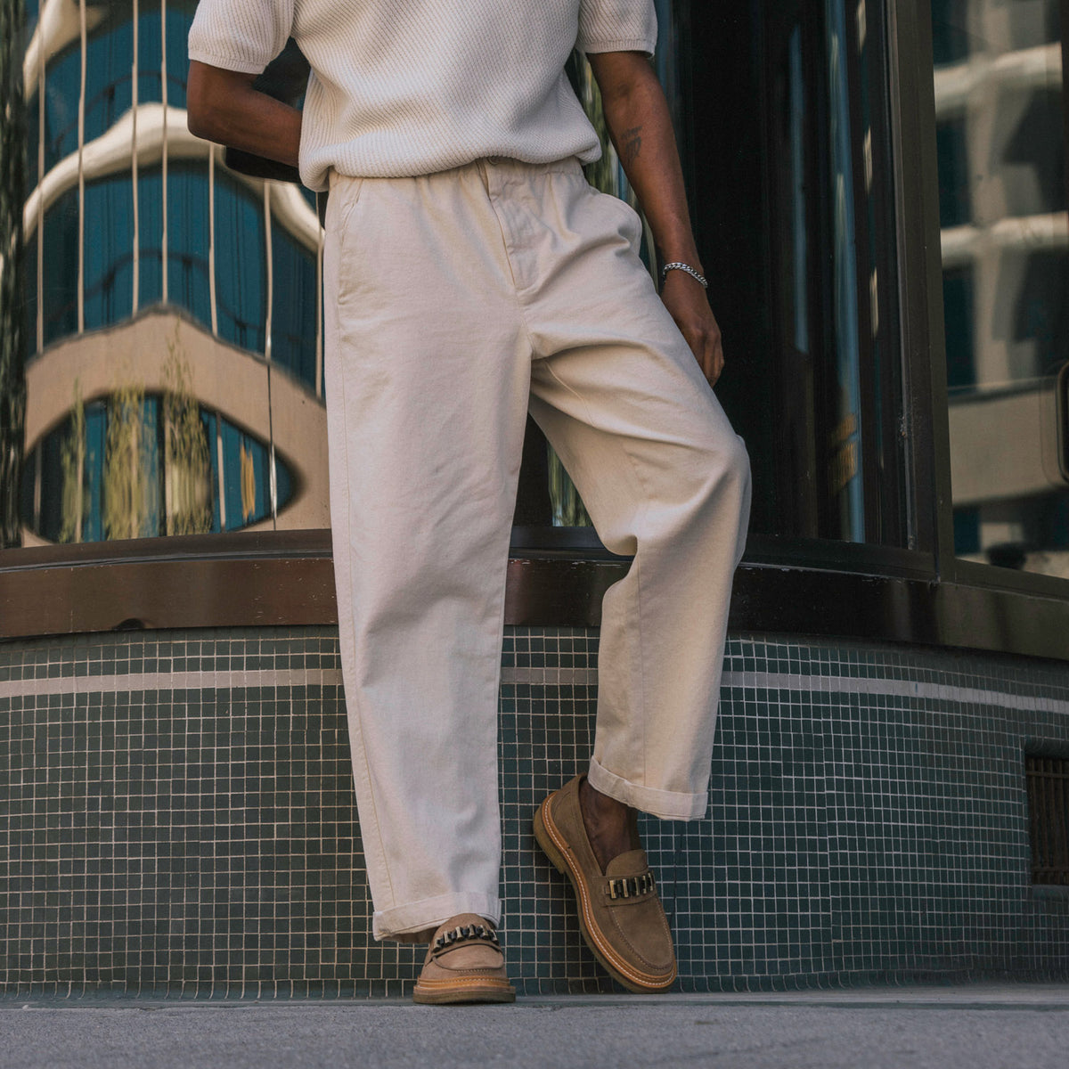 A person wearing light beige pants and brown shoes leans against a reflective building facade.