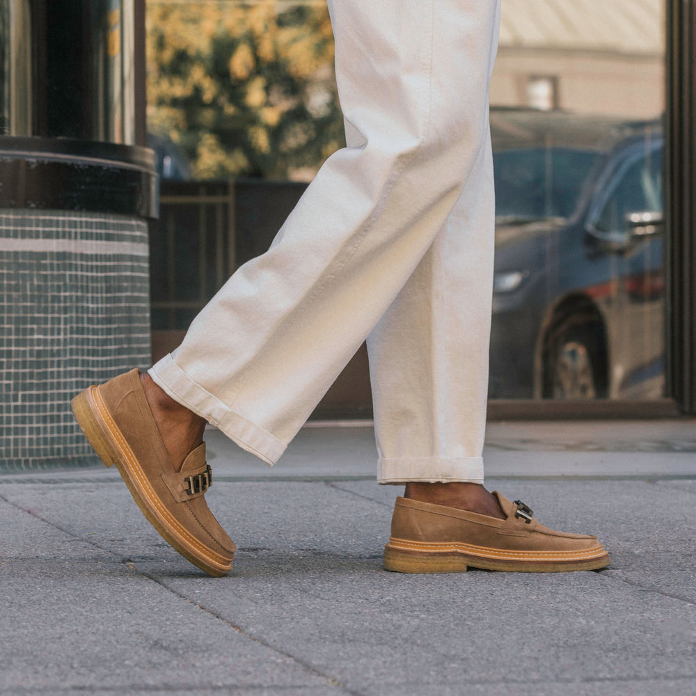 Person wearing white pants and brown loafers walking on a sidewalk.