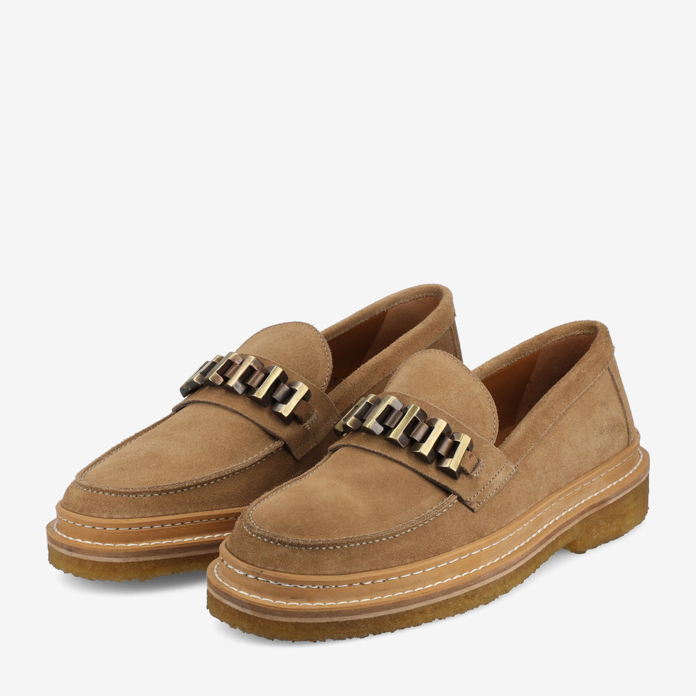 A pair of tan suede loafers with chunky chain embellishments on the upper, thick rubber soles, and white stitching accents.