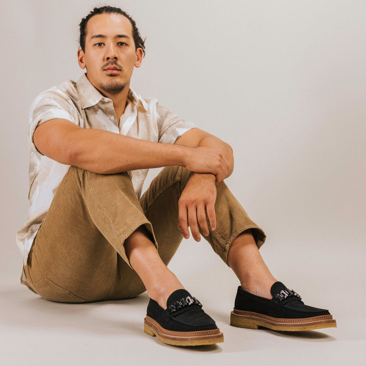 A seated person wearing a short-sleeve patterned shirt, tan pants, and black loafers looks at the camera against a plain background.