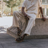 A person in light-colored clothing is seated on a concrete structure, wearing dark brown shoes with metal chain detailing.