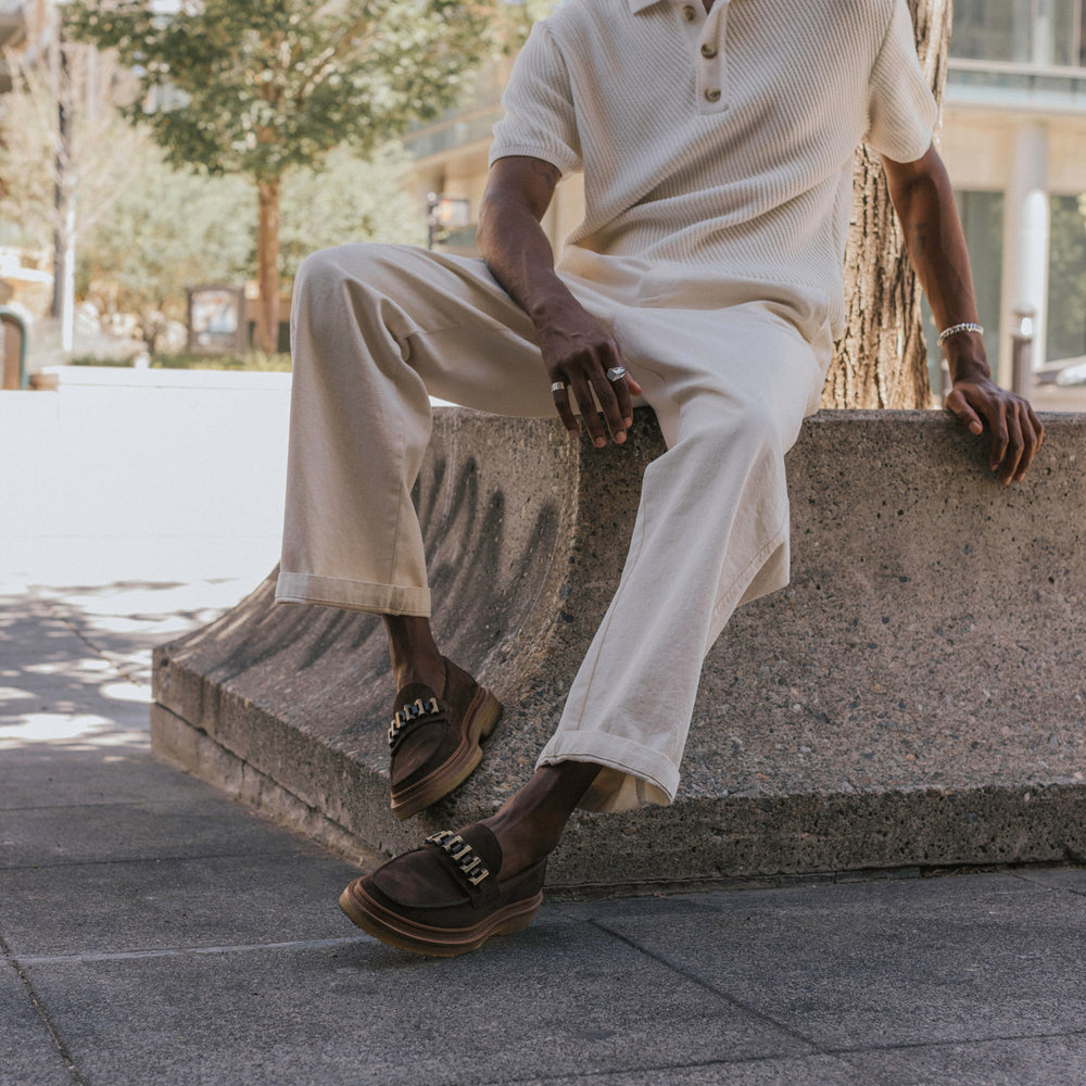 A person in light-colored clothing is seated on a concrete structure, wearing dark brown shoes with metal chain detailing.