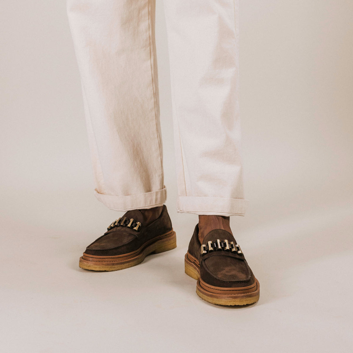 Cropped view of a person wearing white pants and brown loafers with light-colored soles.