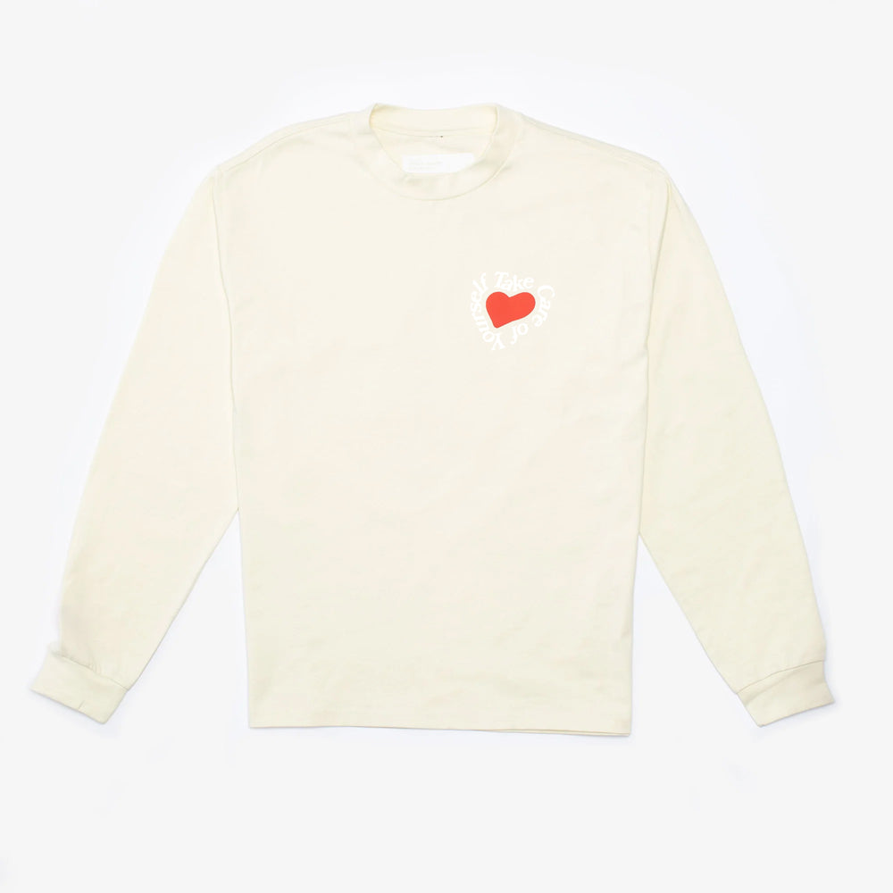 A long-sleeved, cream-colored shirt with a small red heart graphic and white text encircling it on the left chest area, laid flat.