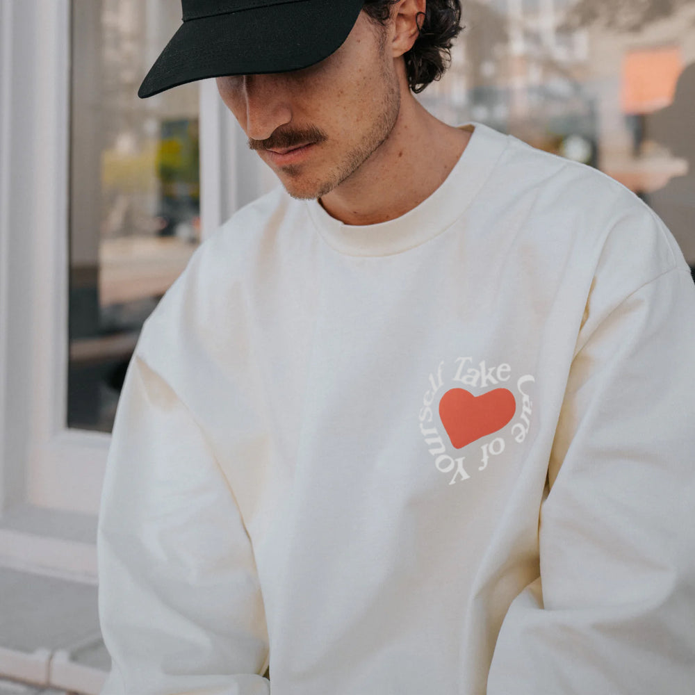 A person wearing a white sweatshirt with Take Care of Yourself written around a red heart shape on the chest and a black cap.