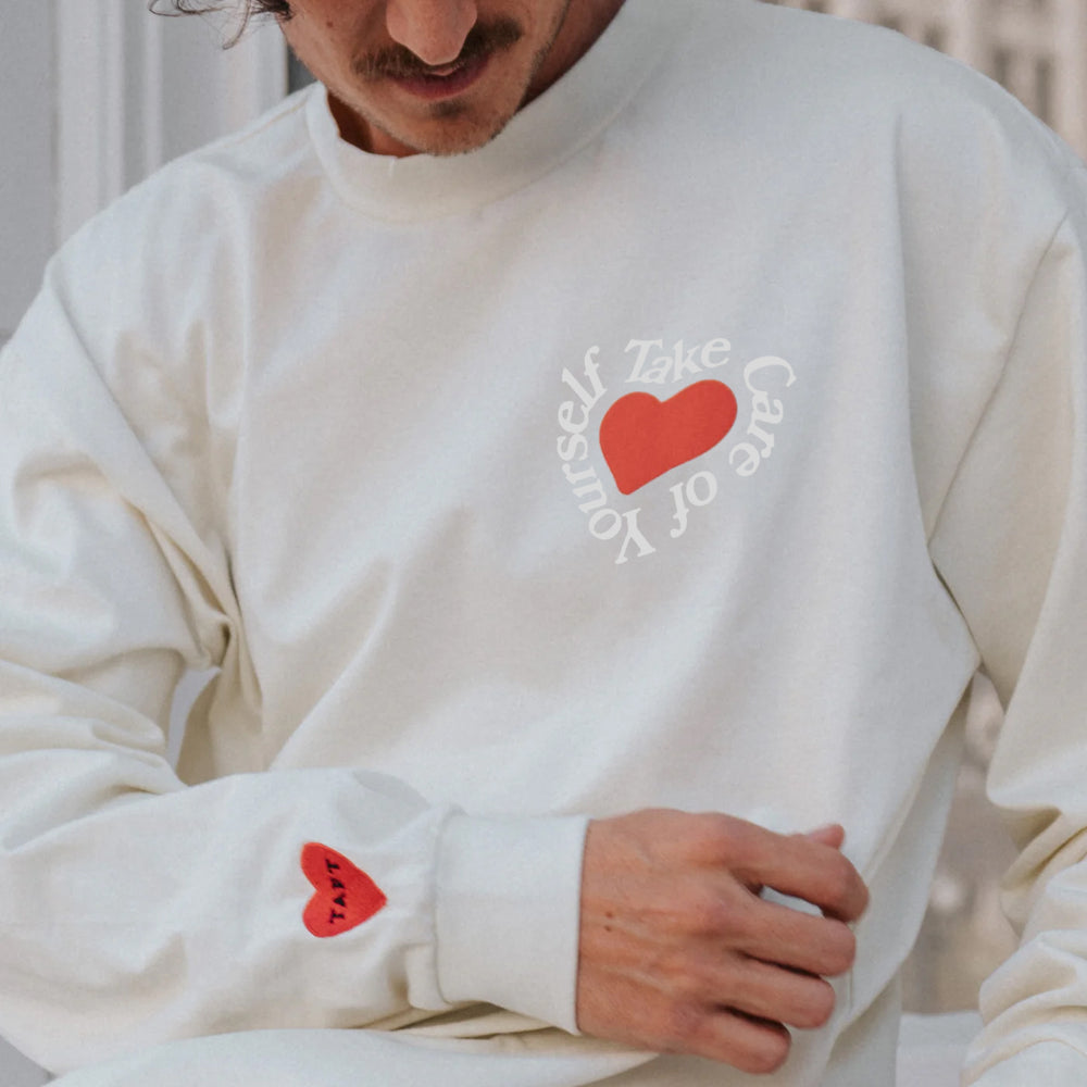 A person wearing a white sweatshirt with a heart graphic and the text Take Care of Yourself printed in a circular design on the chest. Another red heart is visible on the sleeve.