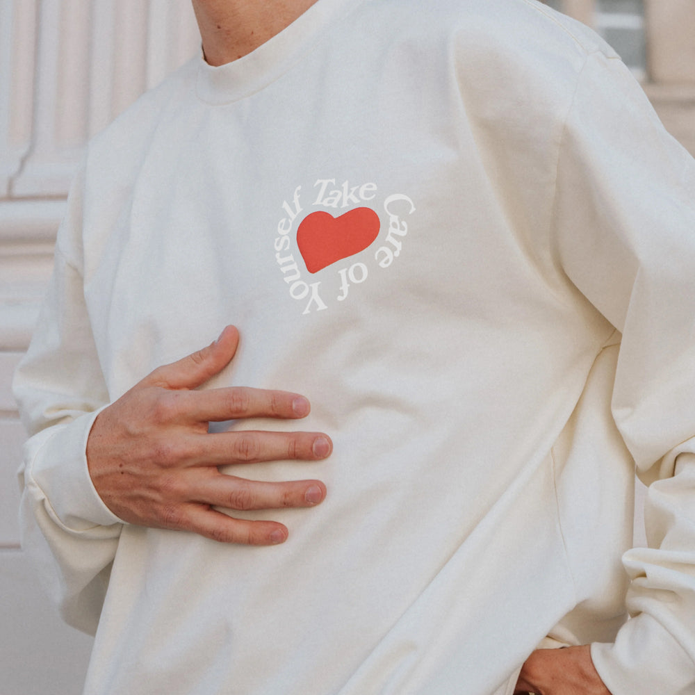 Person wearing a cream-colored sweatshirt with a red heart and the words Take Care of Yourself printed around it.