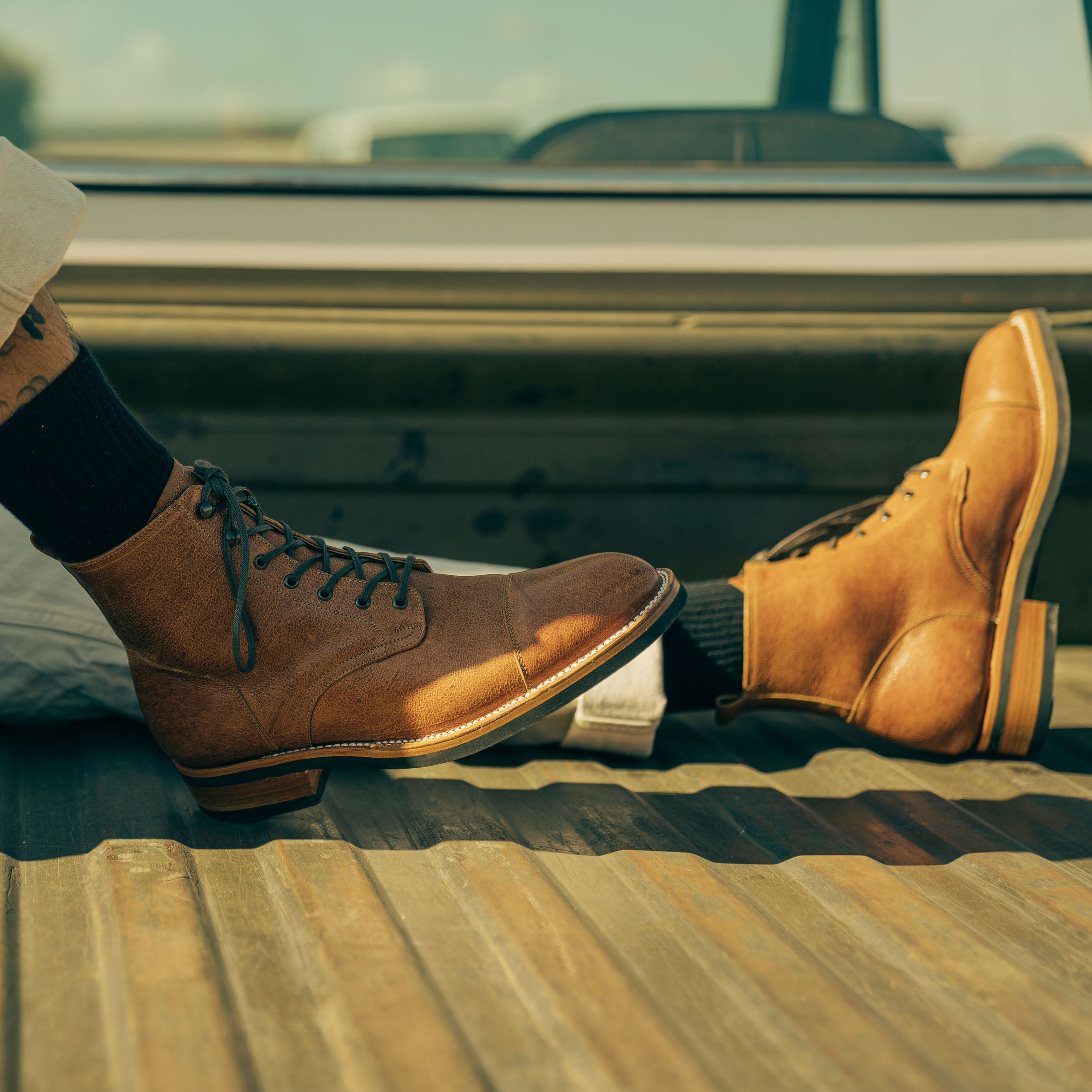 A person wearing tan leather lace-up boots with black socks is sitting in the back of a truck, with legs outstretched and crossed at the ankles.