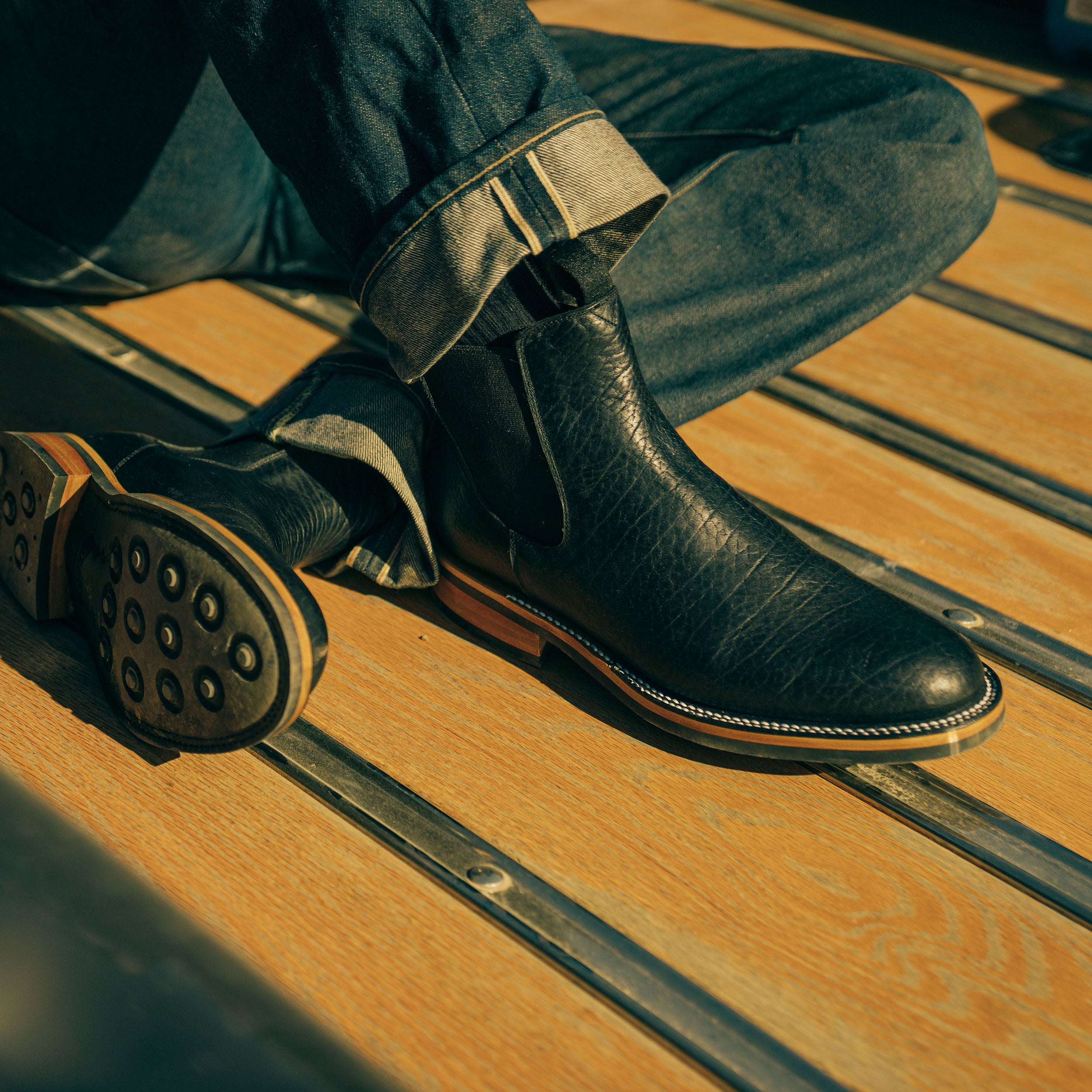 Close-up of a person wearing black textured leather boots and rolled-up jeans, seated on a wooden deck.