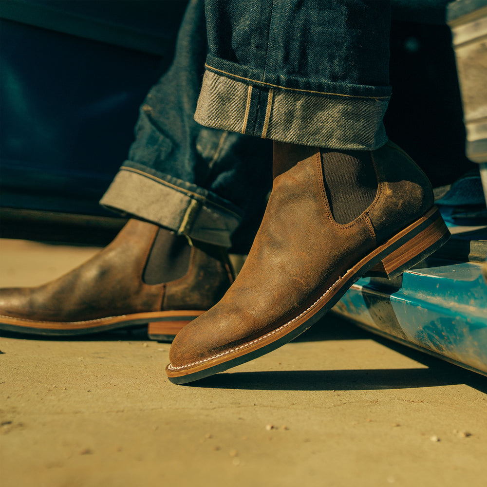 A person wearing brown leather Chelsea boots and rolled-up blue jeans steps out of a blue vehicle onto a sandy surface.