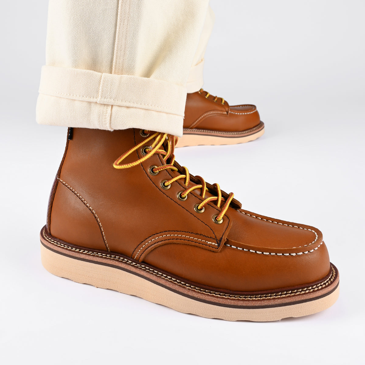 Close-up of a person wearing light-colored pants and brown leather work boots with yellow shoelaces against a plain background.