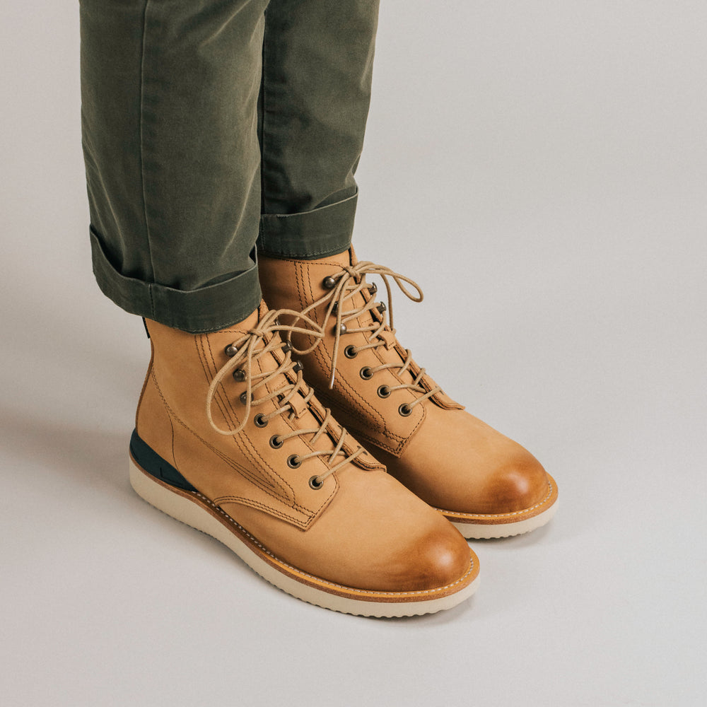 Person wearing green pants and tan lace-up boots standing on a light-colored surface.