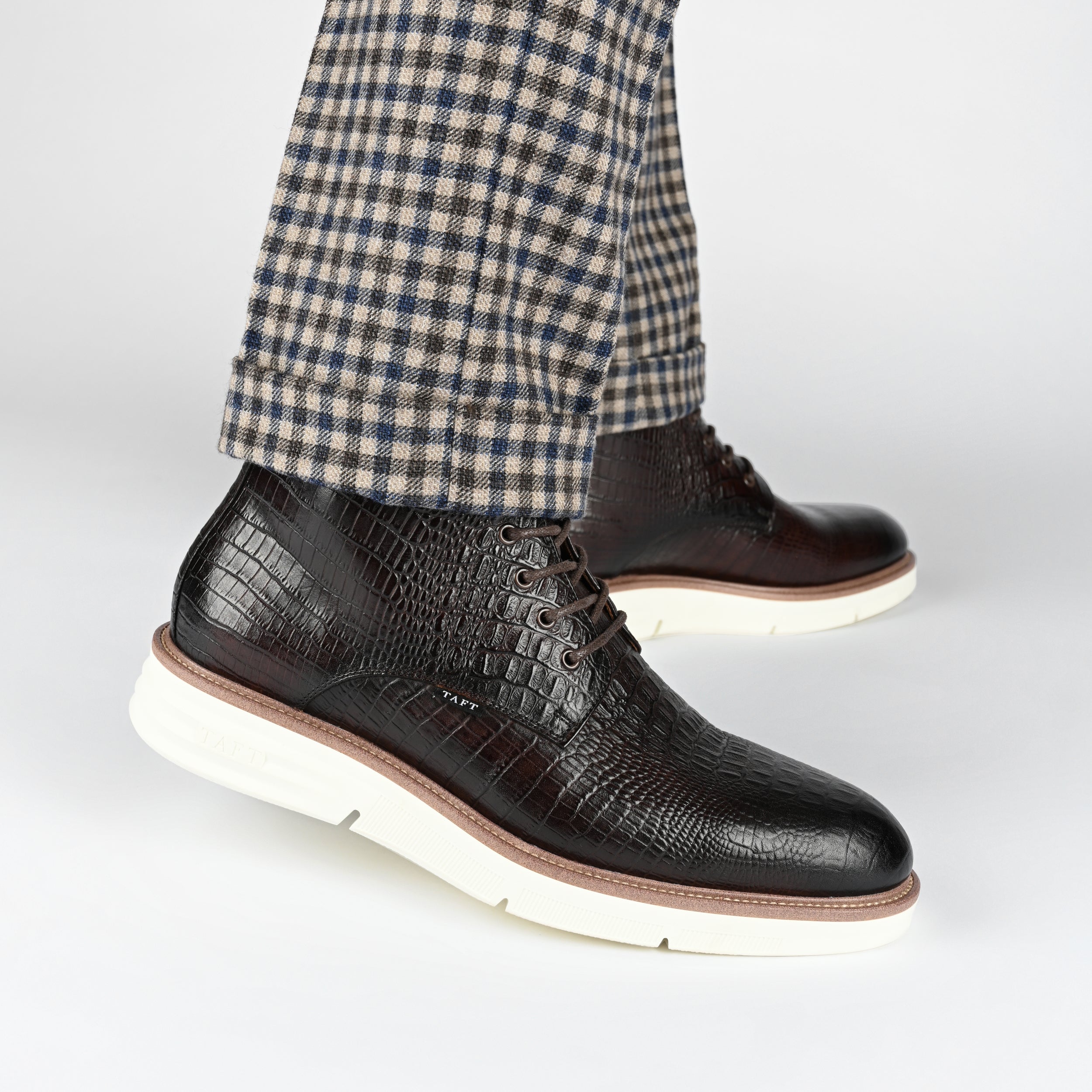 Close-up of a person wearing black textured leather shoes with white soles and checkered brown and blue pants.