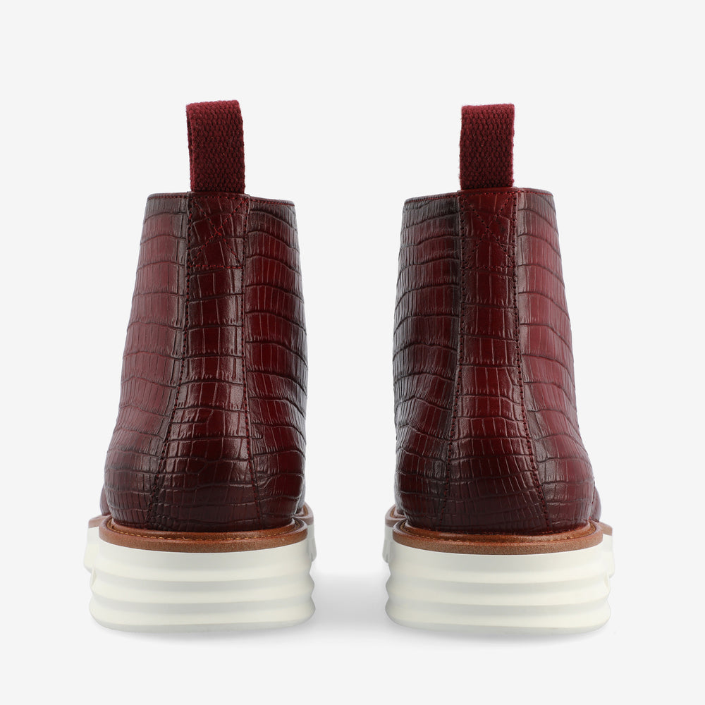 A rear view of a pair of maroon ankle boots with textured crocodile skin pattern and thick white soles.