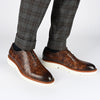 A pair of brown leather dress shoes with white soles, worn by someone in grey plaid pants.