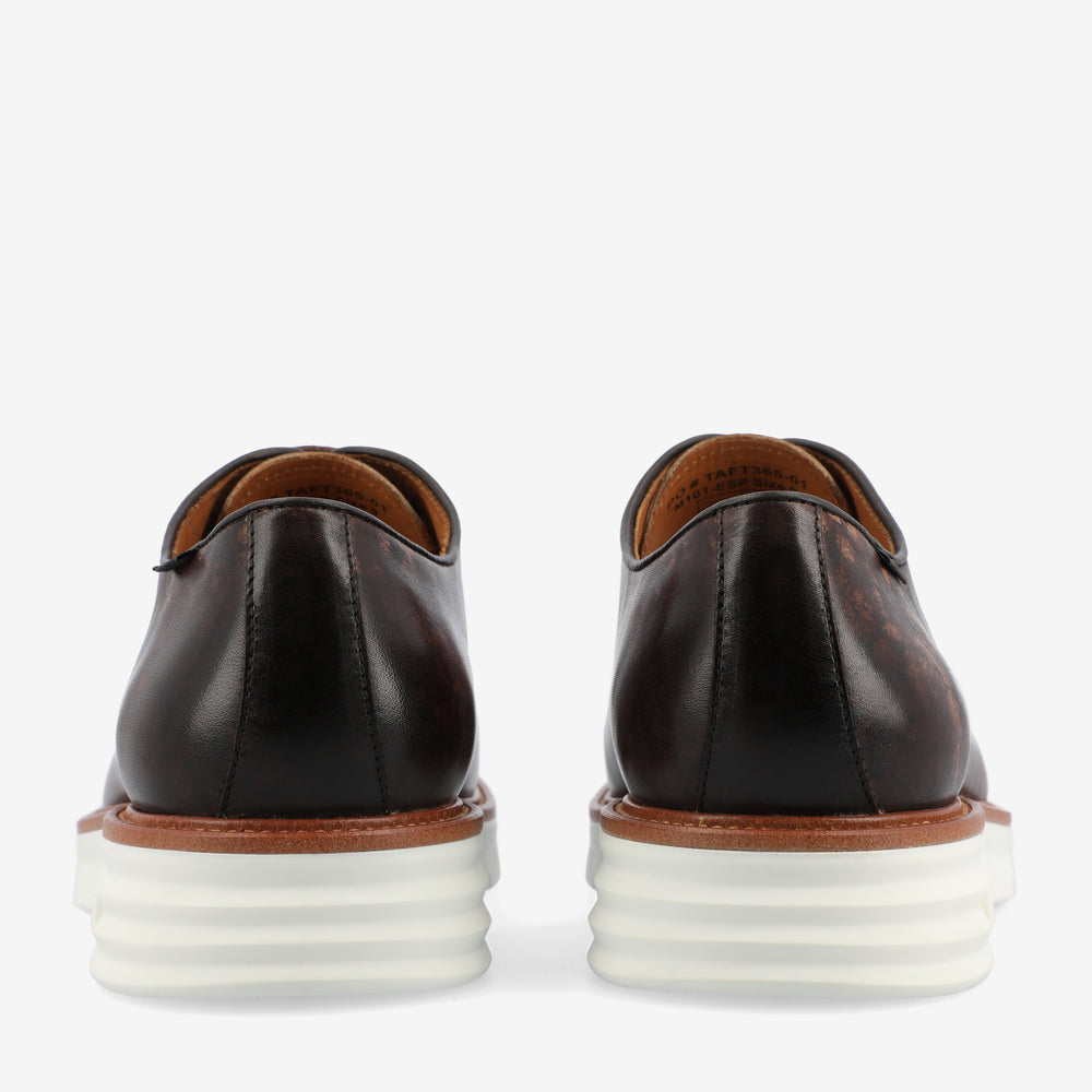 Rear view of a pair of brown leather dress shoes with white stacked rubber soles against a plain background.