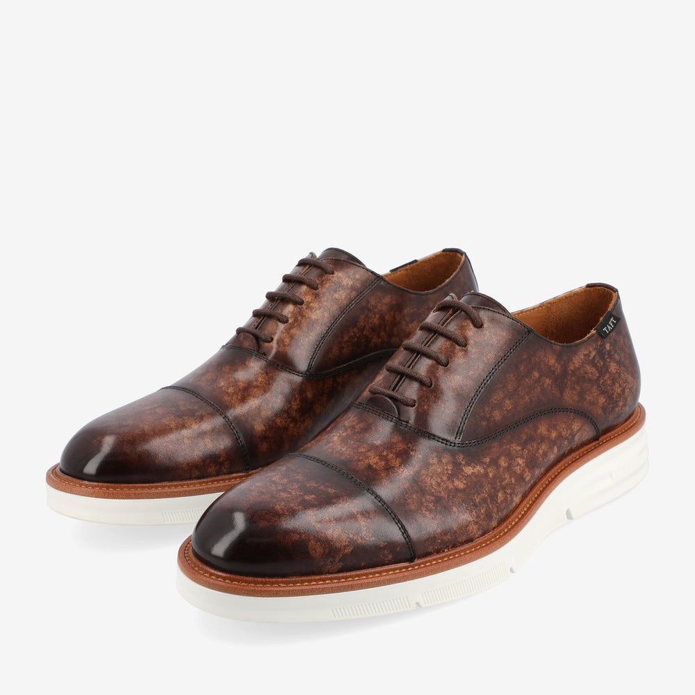 A pair of brown leather dress shoes with white soles and laces, featuring a polished finish and cap toe design, viewed at an angle on a white background.