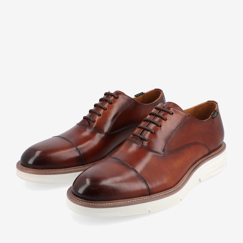 A pair of brown leather dress shoes with white rubber soles and laces, featuring a cap toe design, set against a white background.