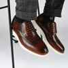 Close-up of a person wearing brown leather brogues with white soles and dark gray pants. The person is seated on a stool, and the background is plain white.