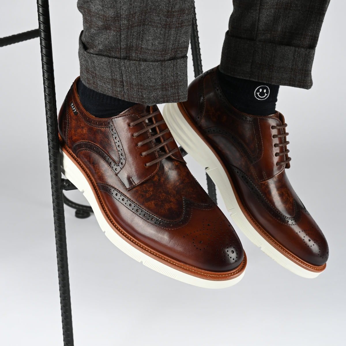 Close-up of a person wearing brown leather brogues with white soles and dark gray pants. The person is seated on a stool, and the background is plain white.