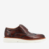 A brown leather wingtip shoe with brogue detailing and white rubber sole, viewed from the side.