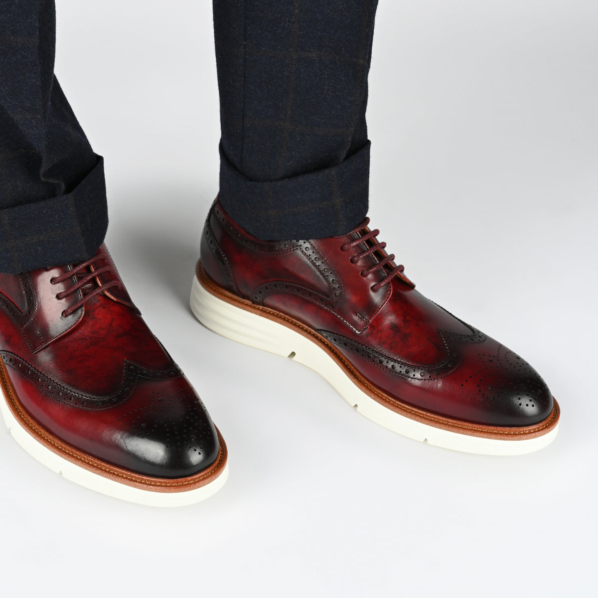 Close-up of a person wearing dark trousers and dark red brogue shoes with white soles standing against a plain background.