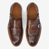 A pair of brown leather monk strap shoes with silver buckles, showing a patina finish and a wingtip design.