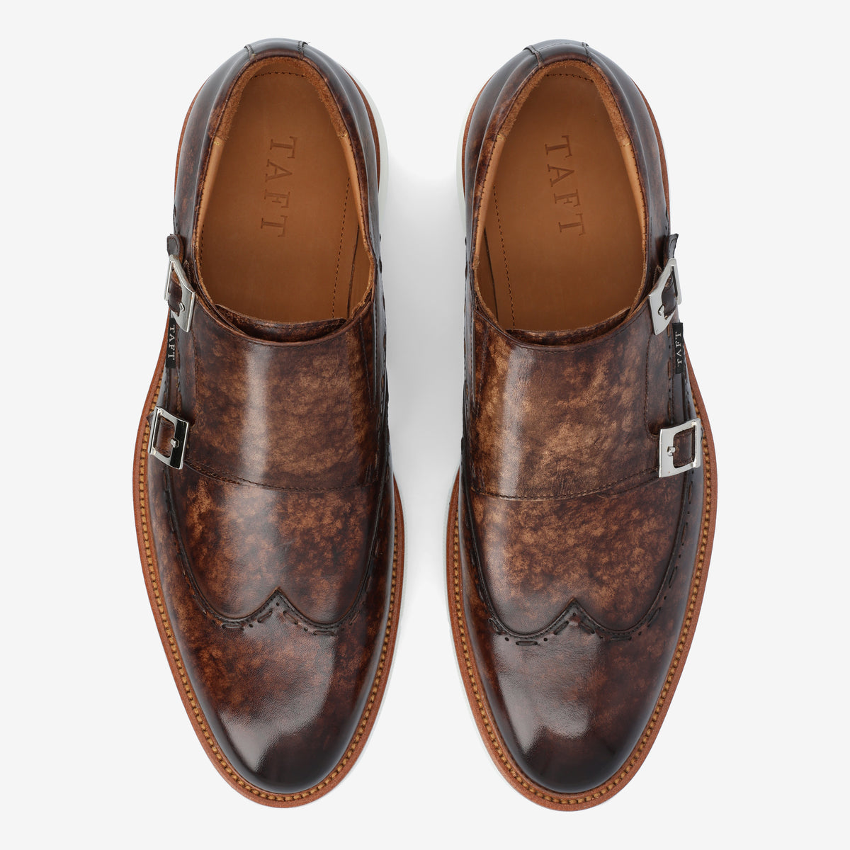 A pair of brown leather monk strap shoes with silver buckles, showing a patina finish and a wingtip design.