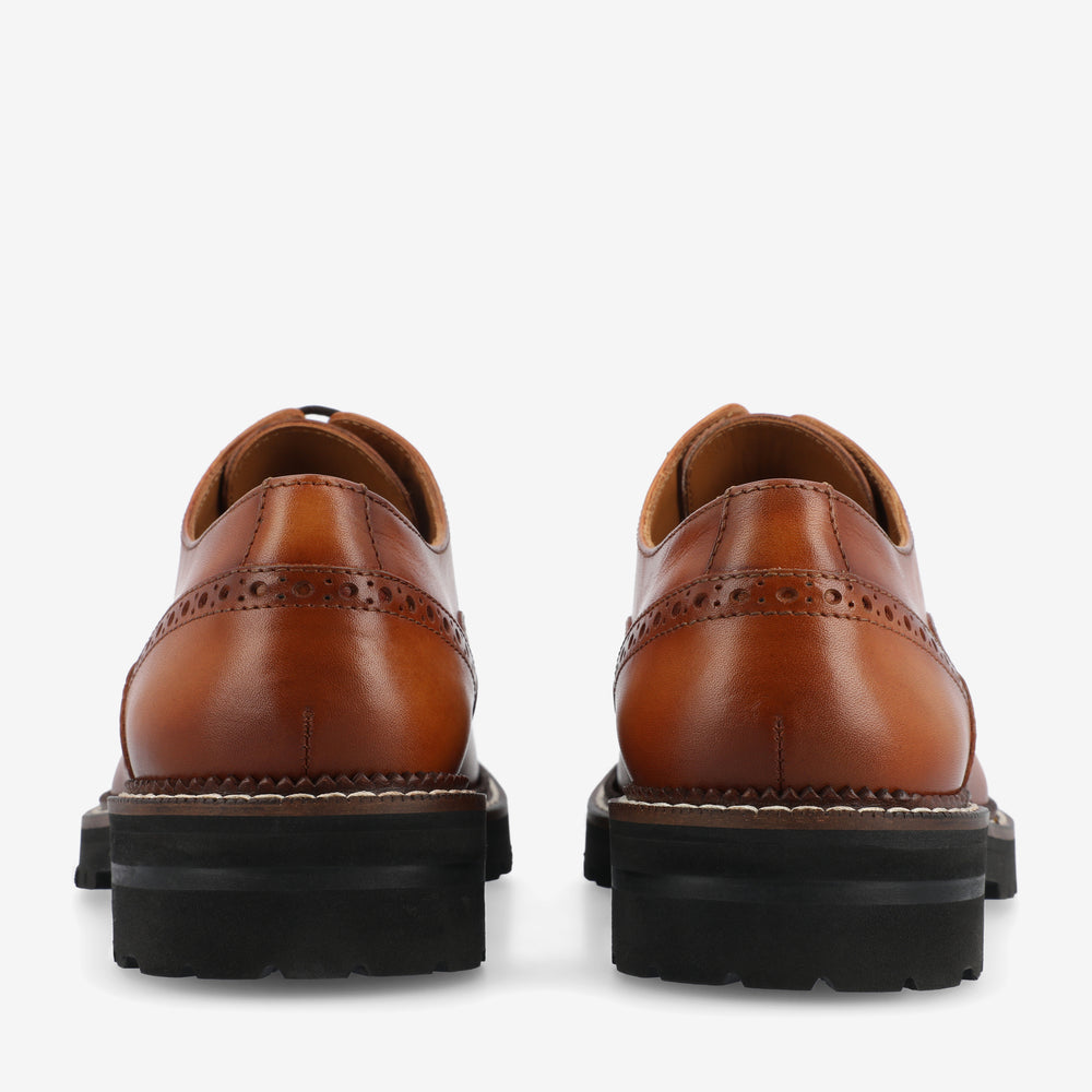 Rear view of a pair of brown leather dress shoes with brogue detailing and black rubber soles.