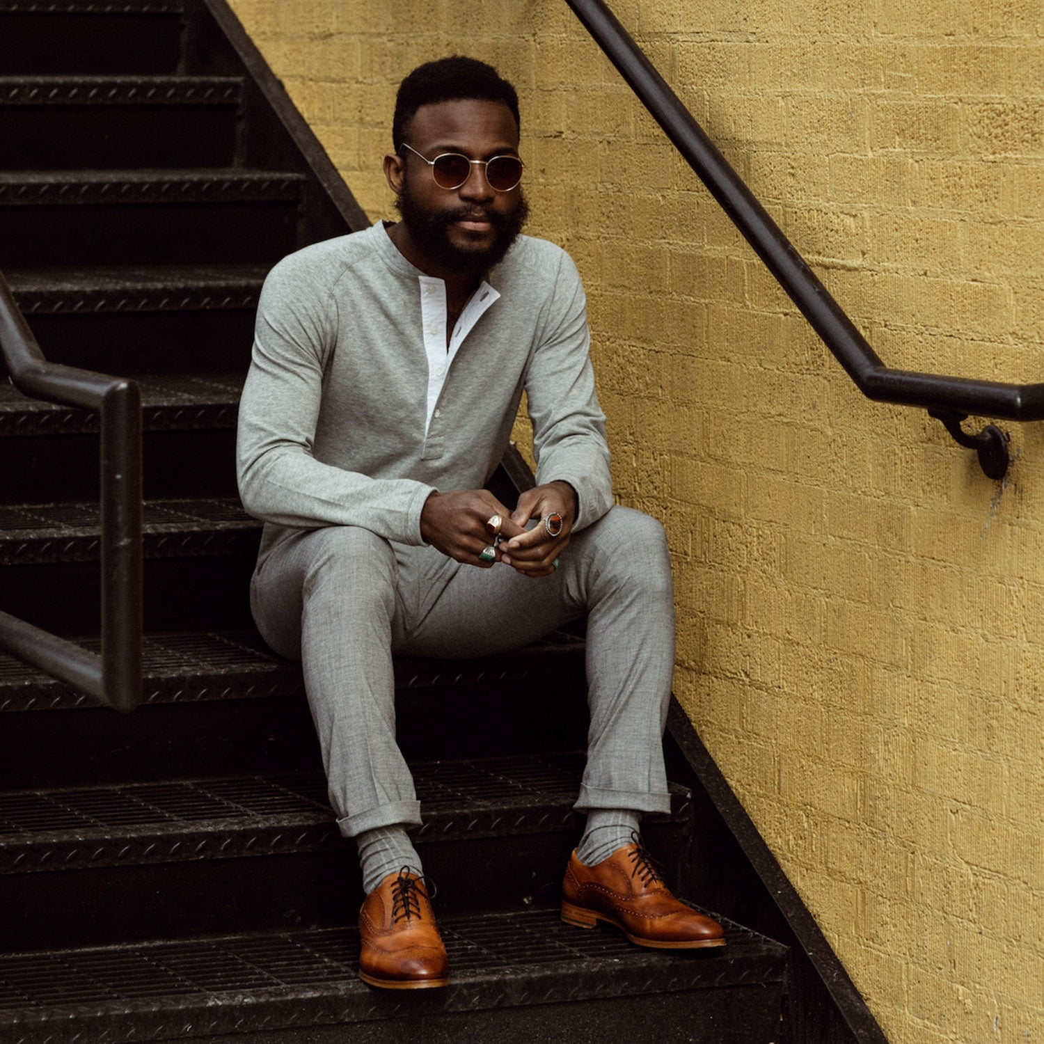 A person with sunglasses and a beard sits on outdoor stairs, wearing a grey long-sleeve shirt, grey pants, and brown dress shoes.