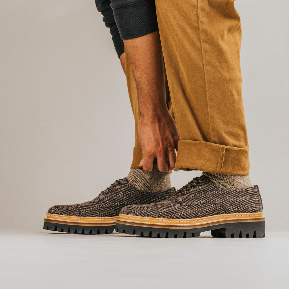 A person wearing mustard-colored pants and gray socks adjusts the cuff of their pants, revealing textured brown lace-up shoes with thick, rugged soles.