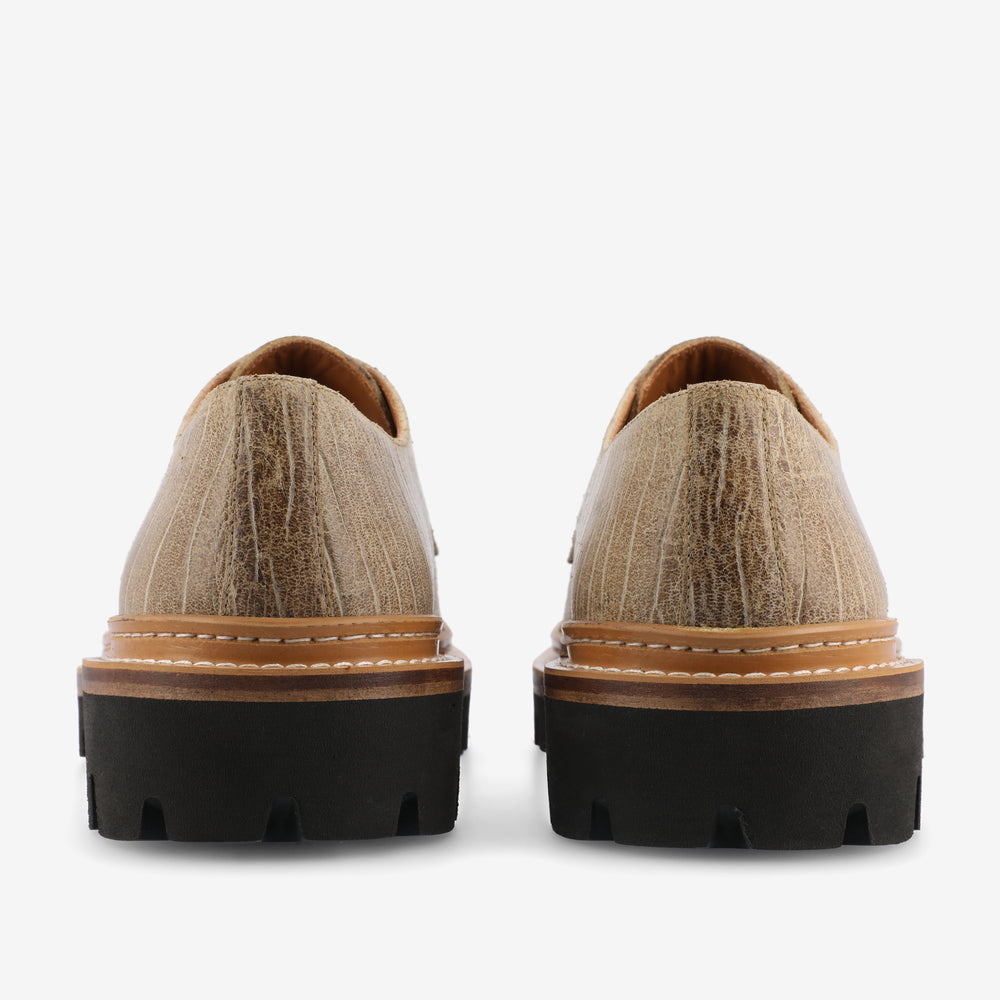 Rear view of a pair of light brown textured leather shoes with thick black rubber soles and white stitching.