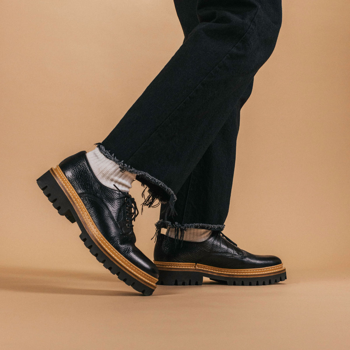 Person wearing frayed black jeans, white socks, and black leather shoes with thick soles, standing on a beige background.