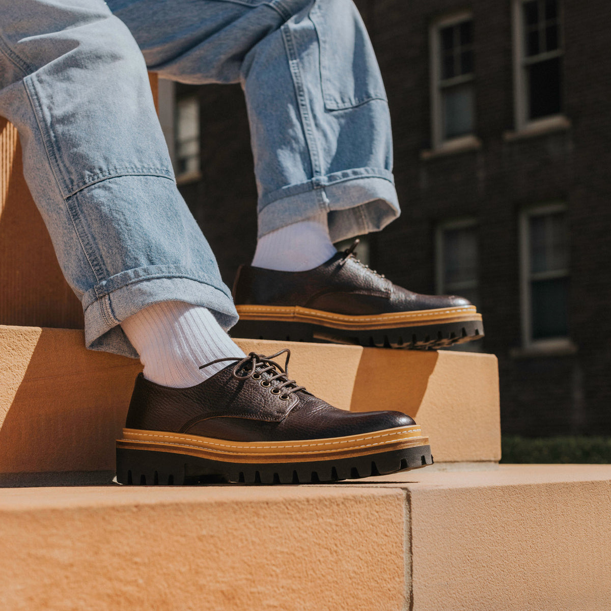 A person wearing blue jeans, white socks, and black laced shoes with thick soles sits on outdoor steps made of beige stone.