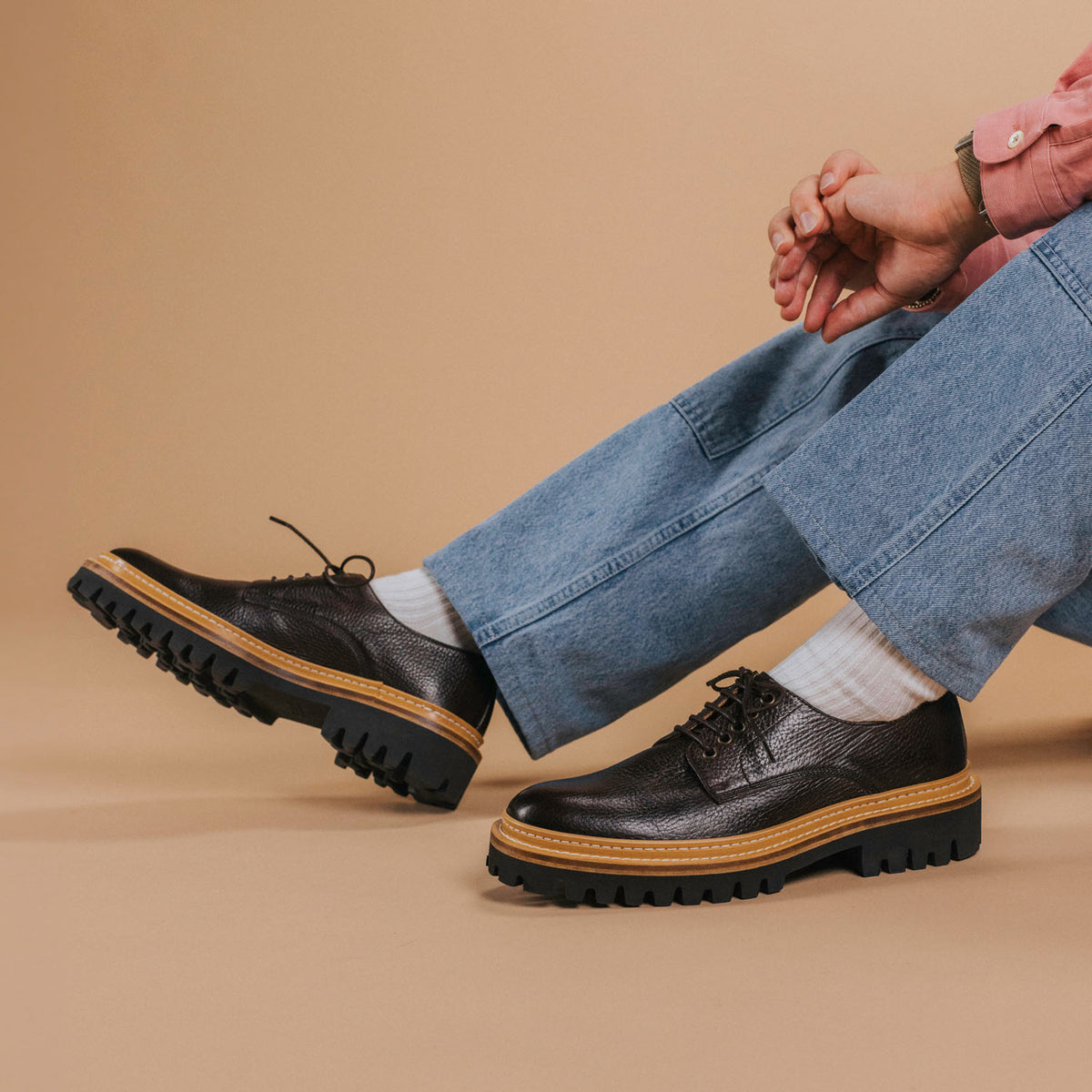 A person in blue jeans and a pink shirt sits against a beige background, showcasing black leather shoes with thick, rugged soles.