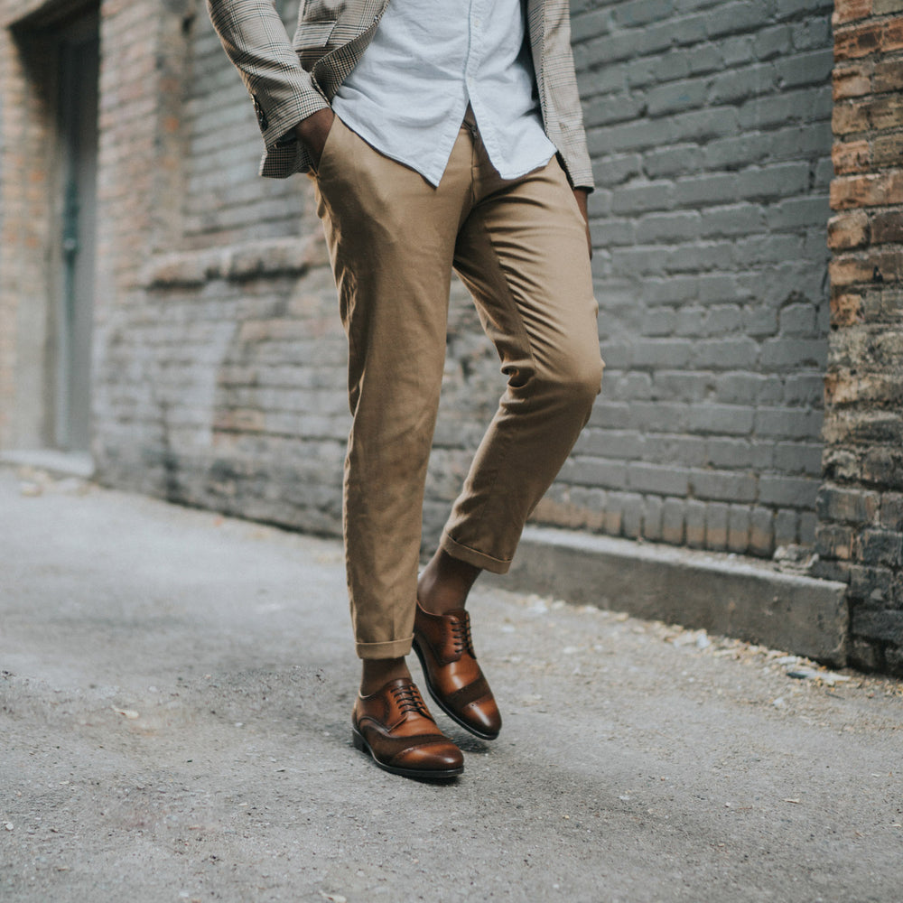 A person wearing brown dress shoes, tan pants, a light shirt, and a checkered blazer stands in front of a brick wall with hands in pockets.