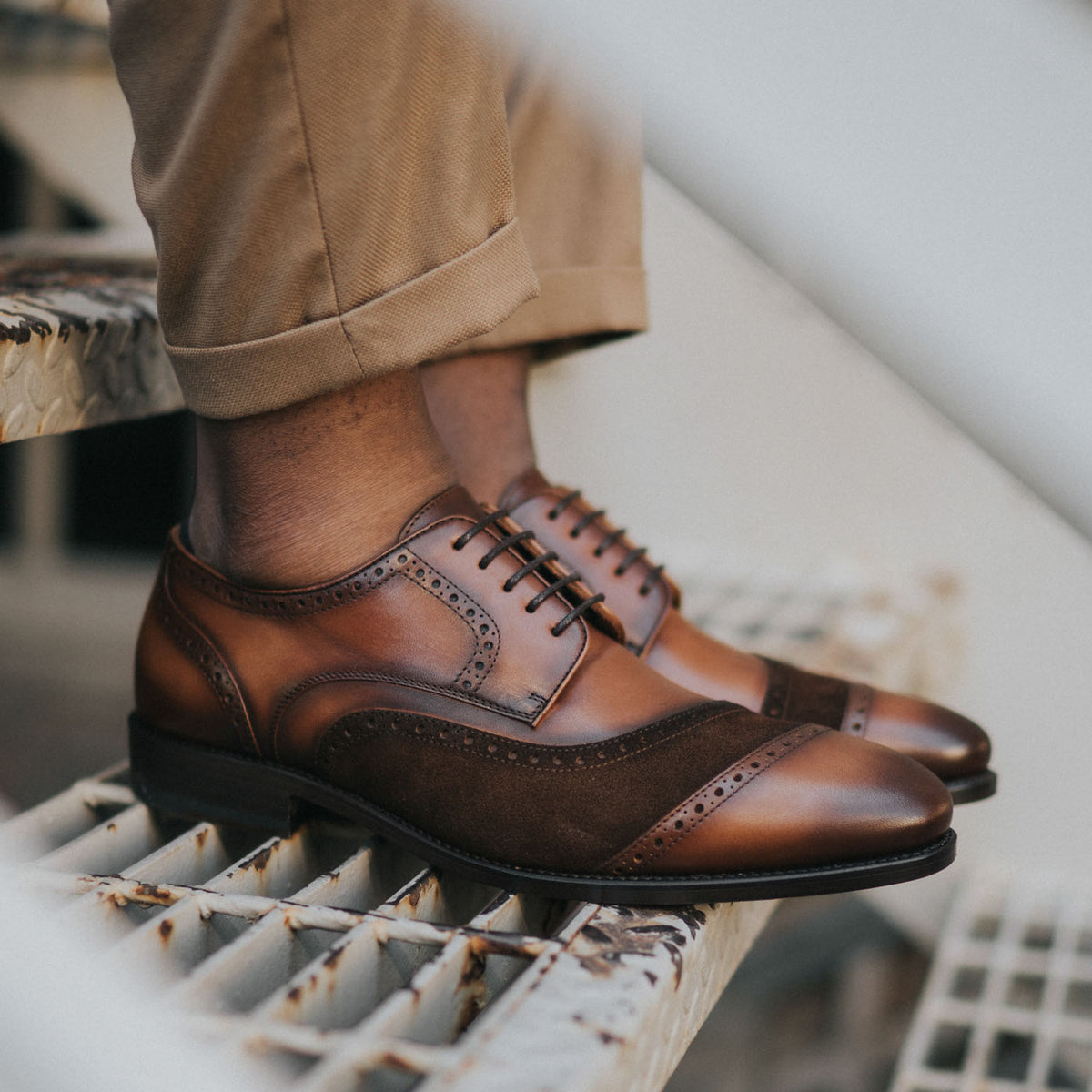 Person wearing brown leather dress shoes with dark suede accents, seated on a metal stairway, wearing beige pants.