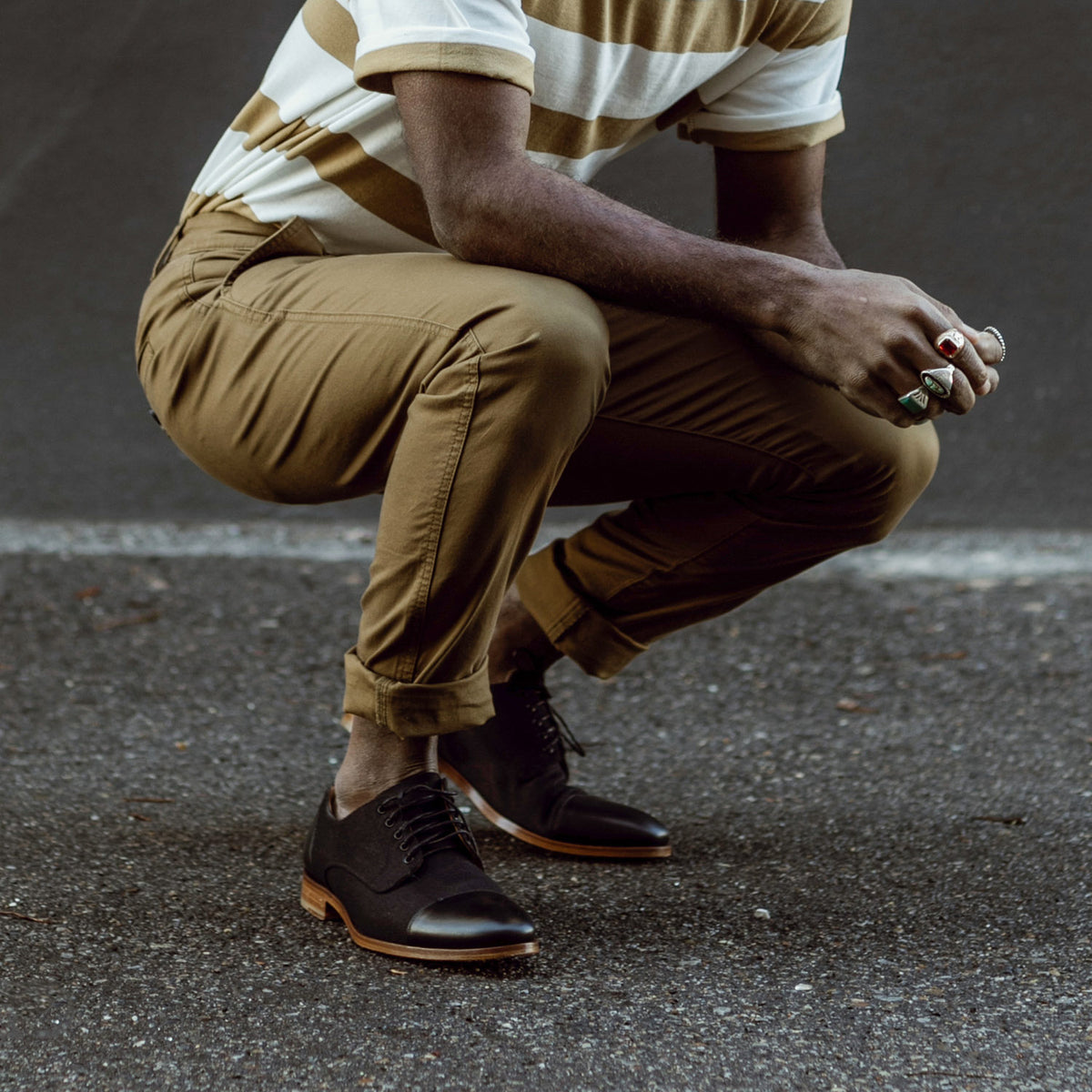 A person wearing a striped shirt, rolled-up khaki pants, and black dress shoes crouches on a paved surface.