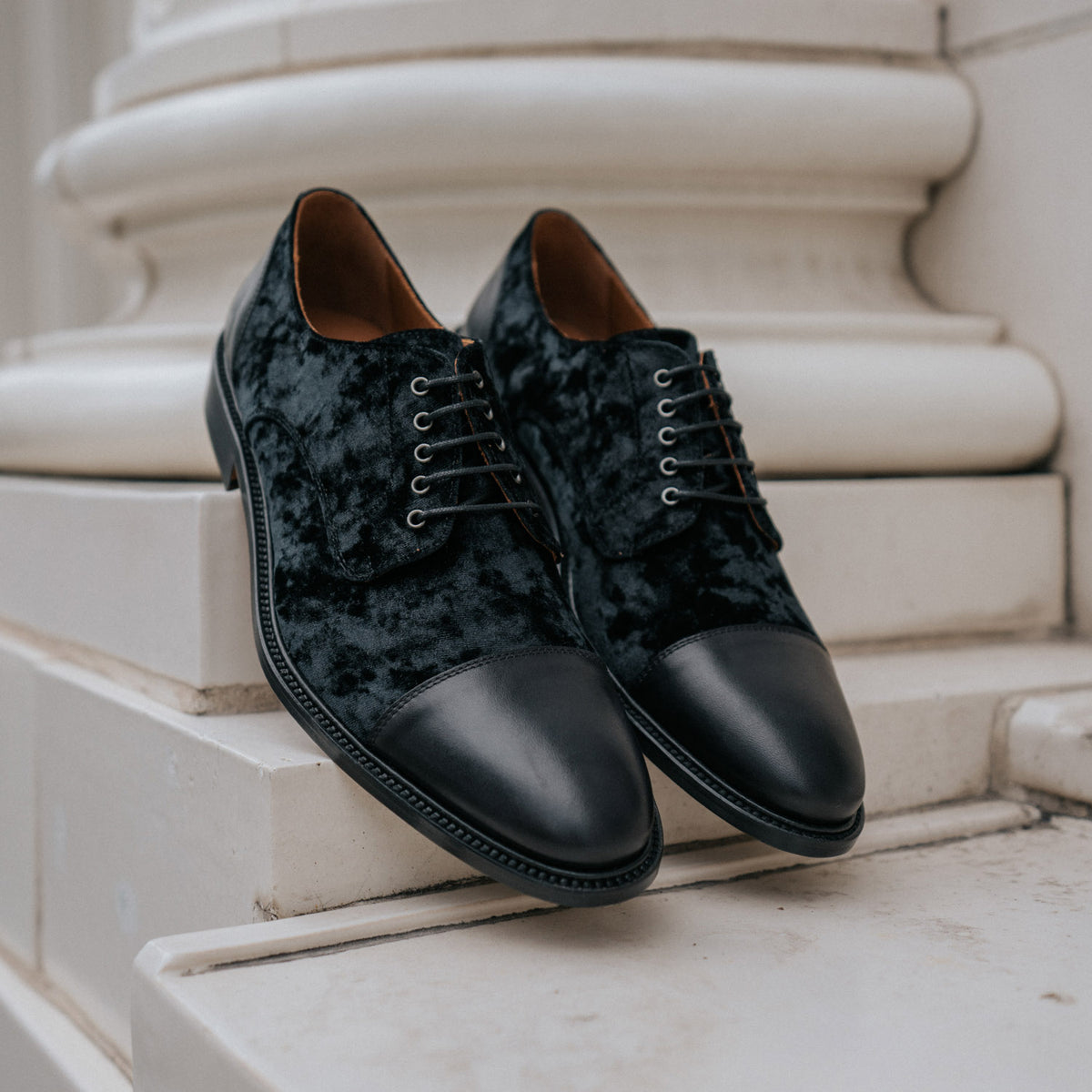 A pair of black velvet dress shoes with leather toe caps and laces, positioned on a white stone ledge.