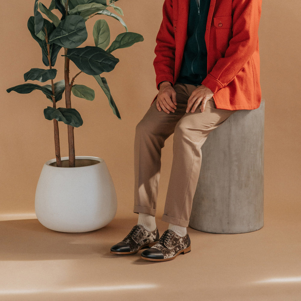 A seated person wearing beige pants, a red jacket, and patterned shoes, beside a potted plant, set against a beige background.