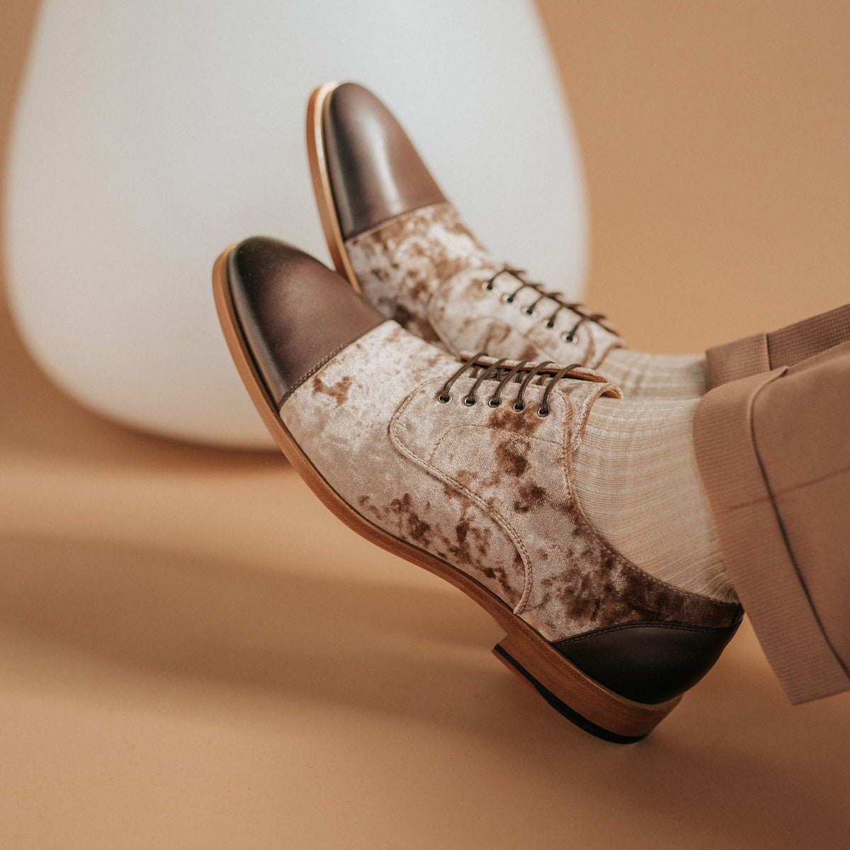 A person wearing beige velvet oxford shoes with brown leather toe caps and white ribbed socks, sits with crossed legs on a beige background.